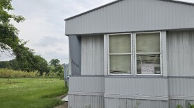 A mobile home sits in a green field with a "for sale" sign in its window.
