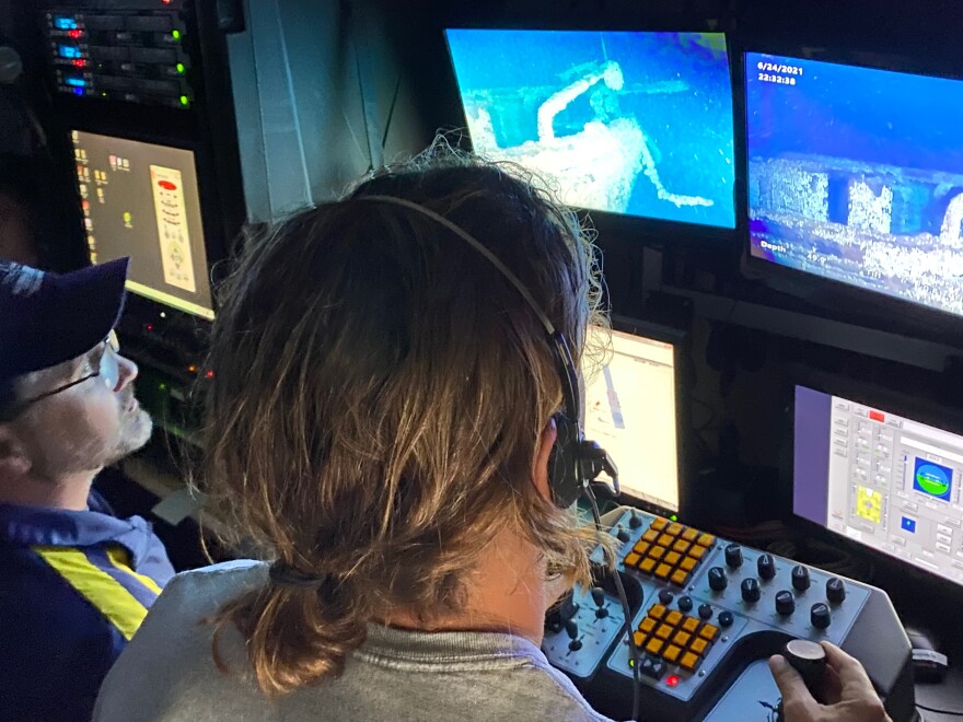 Two people sit at computers and monitors with underwater images of shipwrecks on the screen