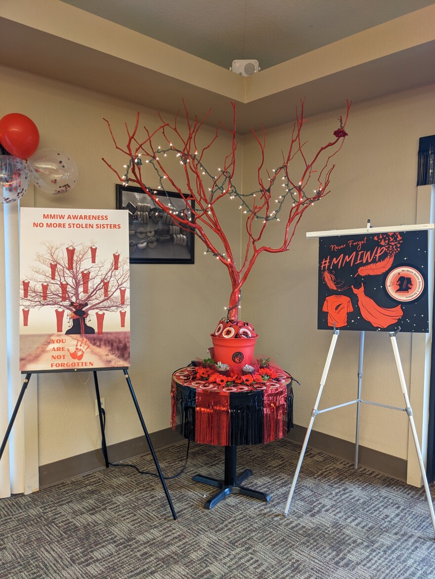 A small red tree with no leaves, on a little table with black and red decoration, between posters with information about Missing and Murdered Indigenous people.