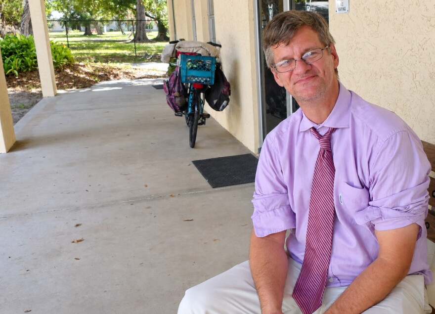 a man with glasses, average length brown hair , violet dress-shirt sleeves rolled up and purple-striped tie sitting on a bench next to a building. behind him leaning on the building is a bicycle 