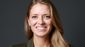 A smiling woman with long blonde hair wears a white shirt under an army green blazer.