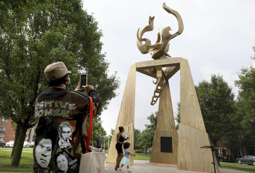 A person wearing a shirt that has images of historical Black women takes a picture of a large sculpture with her cell phone. The sculpture had three pillars leading to an abstract flame and is in honor of Ida B. Wells.