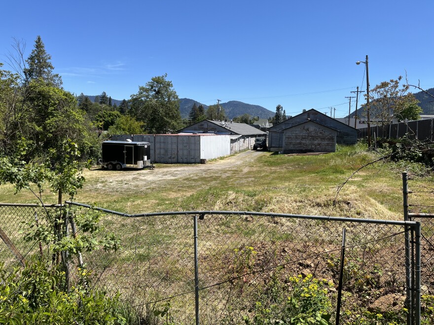 Behind a chain link fence, a green lawn is shown. In the background are buildings. 