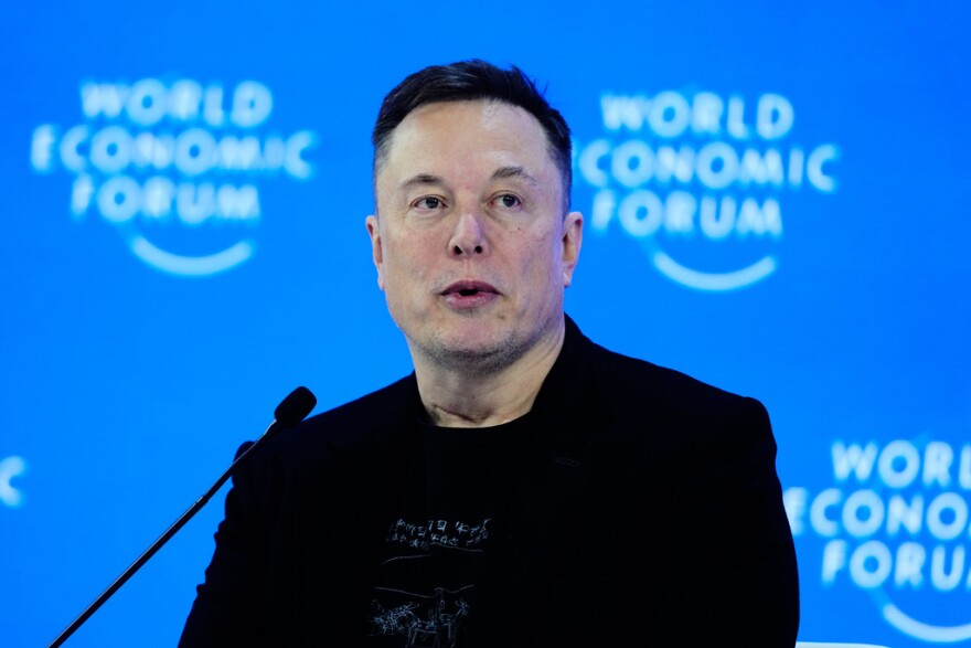 Elon Musk wearing a black jacket sits behind a microphone in front of a blue wall.