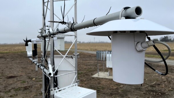 Weather monitoring gadgets are attached to a metal framework. The structure stands in the middle of a farm field.