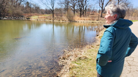 A photo of an older woman in a teal jacket overlooking a small pond. 
