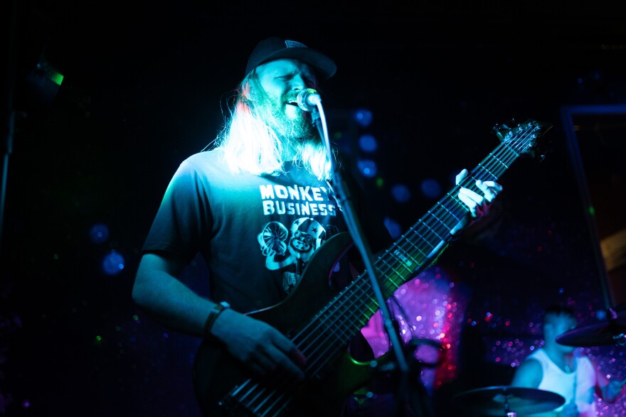 A man in a t-shirt that says "Monkey Business" on the front with a picture of a monkey plays a guitar and sings into a microphone.