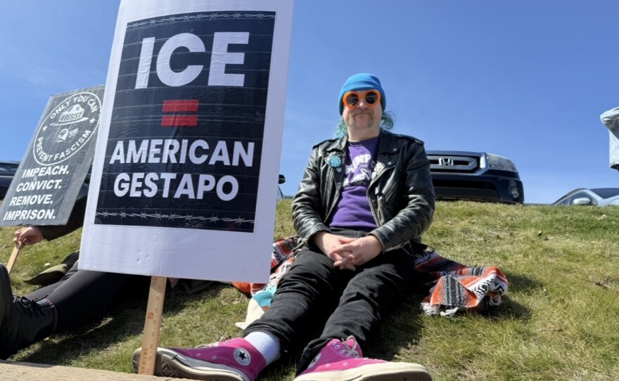 A protester wearing orange flower-shaped sunglasses sits on a grassy hill next to a sign that says "ICE = American Gestapo"