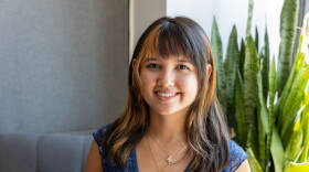A young woman smiles broadly at the camera. She has dark, shoulder-length hair with copper-colored highlights. She wears a septum ring and two pendant necklaces, as well as a lacy blue cap-sleeved top. Behind her, a gray couchback and a plant with long, vertical leaves.