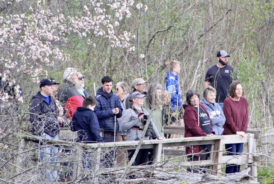 A group of smiling birders stands together outside on a wooden pier to listen for birds on a cloudy spring day. All of the birders are wearing coats, and some are holding white canes to help them navigate the area. 