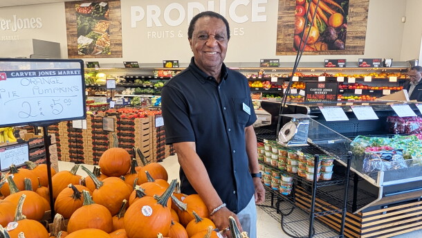A man stands next to a bin of pumpkins that are priced two for $5. The man wears a black, button-down shirt and a name tag. The rest of a grocery store produce department is in the background.