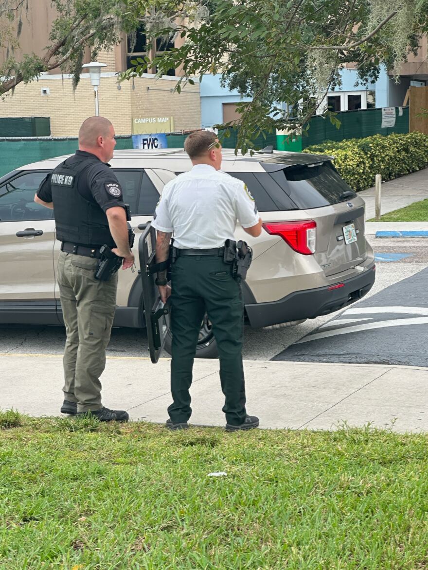 The backs of two law enforcement officers. One holds a rifle in his left hand. A vehicle and building are behind them.