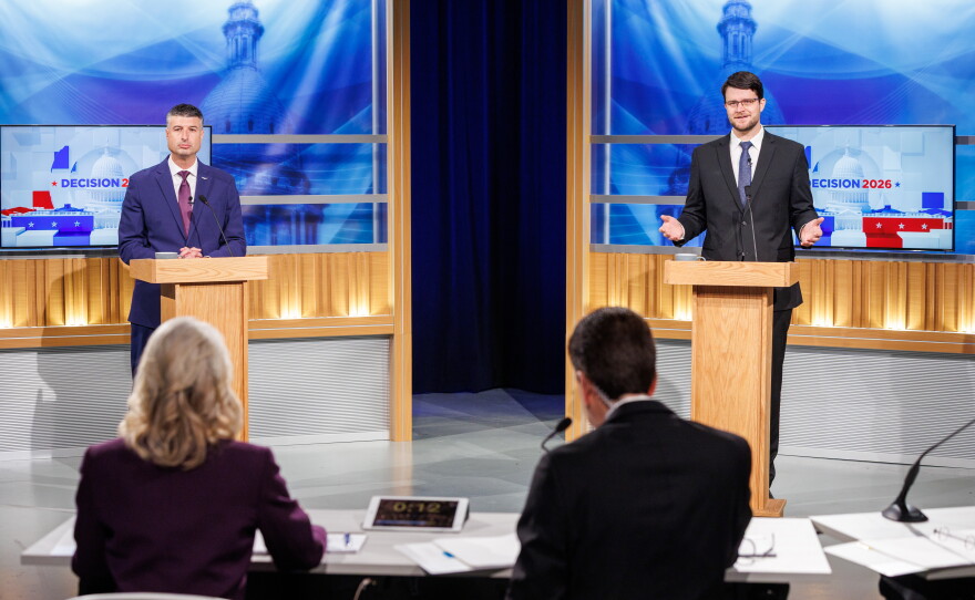 Two men in suits stand at podiums facing a man and woman sitting at a table in a TV studio