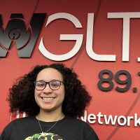 Image of Bloomington High School Student Ameena Gaston standing in front of a red wall with a WGLT logo. She wears glasses, a necklace and a black t-shirt