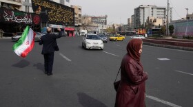 A man waves an Iranian flag during a campaign in support of the government as a woman and vehicles pass by at the Enqelab-e-Eslami, or Islamic Revolution, square in downtown Tehran, Iran, Sunday, March 22, 2026. (AP Photo/Vahid Salemi)