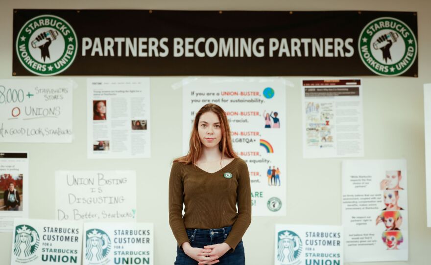 A young woman with long brown hair stands in front of a wall covered in posters.