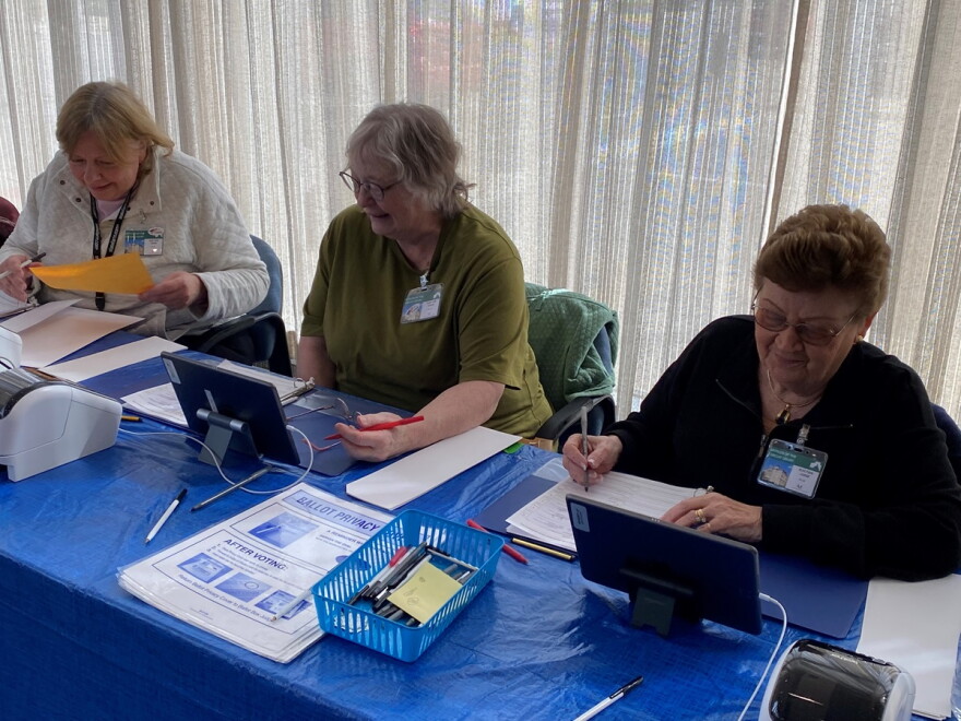 Three women seated at a long table looking at computer tablet screens and writing notes at a polling site