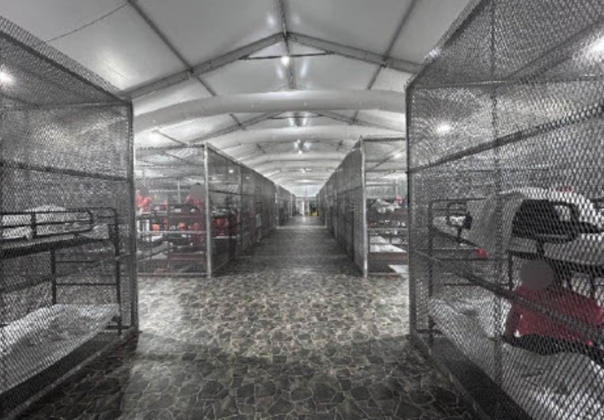 Cages filled with men in orange jumpsuits line the floor inside a tent. 