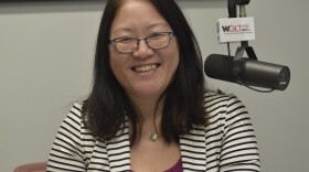 State Rep. Sharon Chung in a radio studio in a black-and-white striped suit jacket seated next to a microphone with the mic flag labeled 'WGLT.org 89.1 FM'