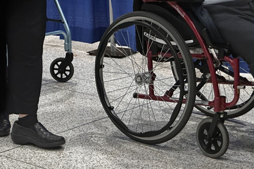 A person using a wheelchair moves along a tiled floor while in line. The wheelchair is red with black and grey wheels and accents. Behind them, a person wearing black pants and socks walks, and someone using a teal walker is behind them. 