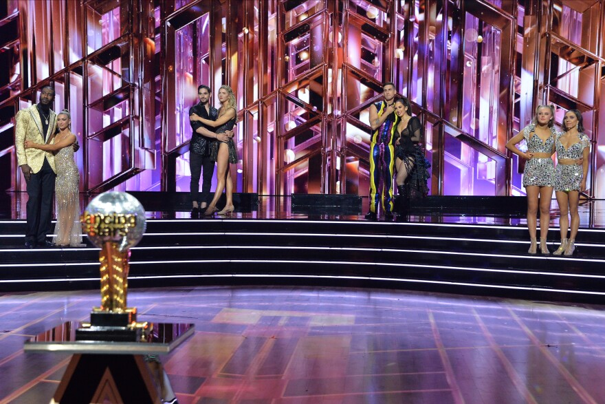 four couples in evening dancewear stand on a tiered stage. a trophy is in the foreground.