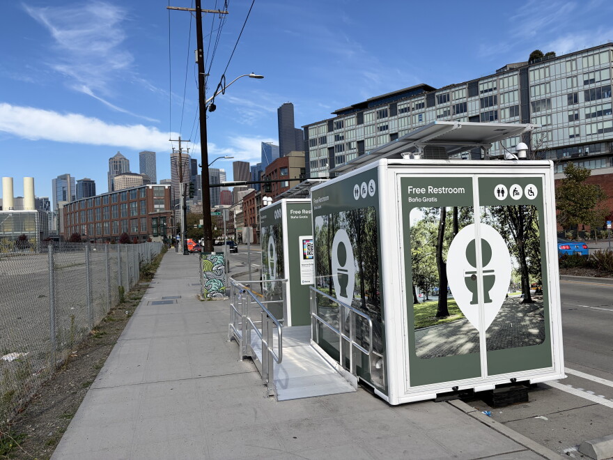 A photo of two Throne bathrooms with the Seattle skyline visible in the background. The bathroom trailers have images of trees on the outside and signage that says "free restroom" and an image of a toilet. 