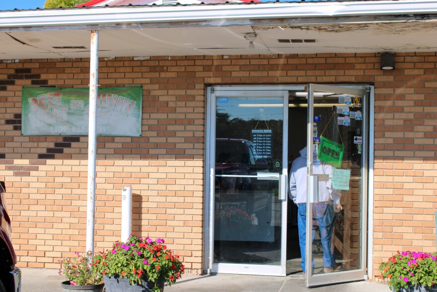 Bloomington Meats entrance and exterior. A metal awning is over the brick building, with glass front doors. A men enters using the right door, where there are some small stickers and a neon green open sign. A sign hanging from the left door has the hours. To the left of the double doors is a faded green sign with red letters spelling "Best meat on Bunn Street" and the store phone number.