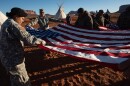 Photo of women and men in military uniform holding open a large american flag, with a tipi in the background. 