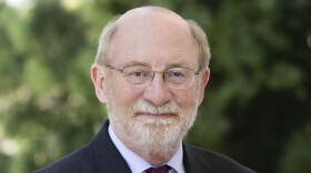 A bald man in a suit and dark purple tie looks into the camera. He is wearing glasses, a pin is on his right lapel. 