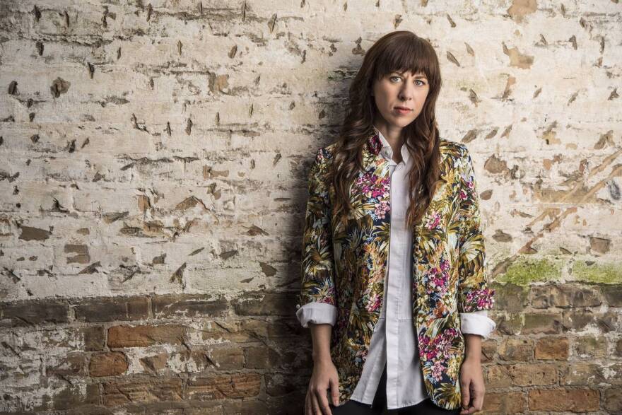 composer Missy Mazzoli stands in front of a rustic brick wall, wearing a pink shirt and floral jacket
