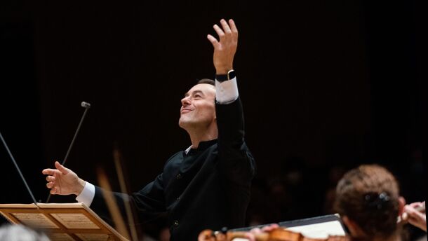 Jonathan Cohen stands at the podium with his hand raised high above him. He gazes upwards and smiles.