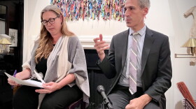 A woman with long blond hair and thick black glasses holds a stack of papers and a pen as she listens to remarks made by a man beside her who has gray hair and wears a gray suit. They are seated on stools in front of a fireplace that is adorned with a colorful artwork made of bits of fabric.