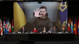 a man appears on a screen behind five people sitting at a long desk surrounded by various countries' flags