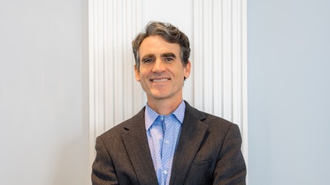 A man in a dark suit jacket and light blue patterned shirt stands smiling in front of a white wall.