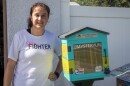 A woman in a white shirt with text reading "she's a fighter" stands next to a small wooden box painted teal and green with a clear door.