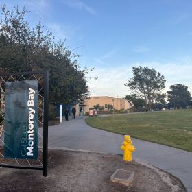 A vertical banner that reads Cal State Monterey Bay stands next to a walking path with a yellow fire hydrant beside it.