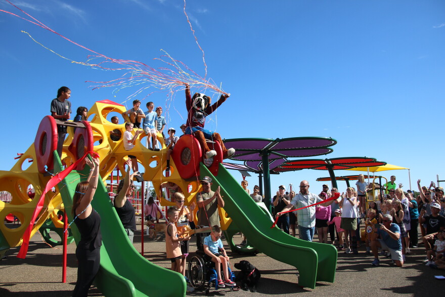 A large crowd of people cheer and clap their hands while looking at a colorful play structure. There's kids on and around the play structure. On top of one of the slides on the structure is the mascot for the Colorado Avalanche releasing confetti. In front of the play structure is Miles Bowling and his family. 