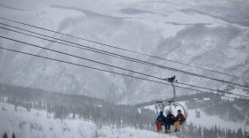 A group of three skiers sits on a chairlift in front of snowy mountains.