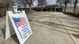 A white sandwich board sign rests on the concrete outside a building. On the sign is an illustration of an American flag, waving. The sign says VOTE EARLY HERE TODAY in blue letters, and 9a - 5p underneath