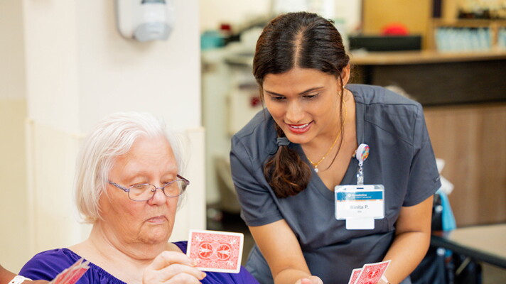 A nurse in scrubs leans over while holding playing cards next to a nursing home resident, also holding playing cards.