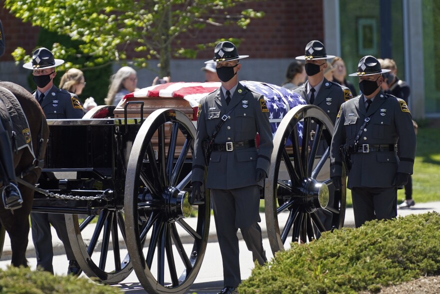 horse pulls casket on buggy during funeral while men in dark uniforms walk alongside