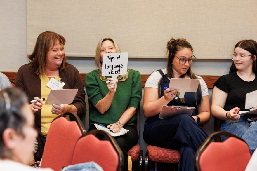 Four women sit next to each other, holding pieces of paper. One woman is smiling as another holds up a card that reads "type of language used". 