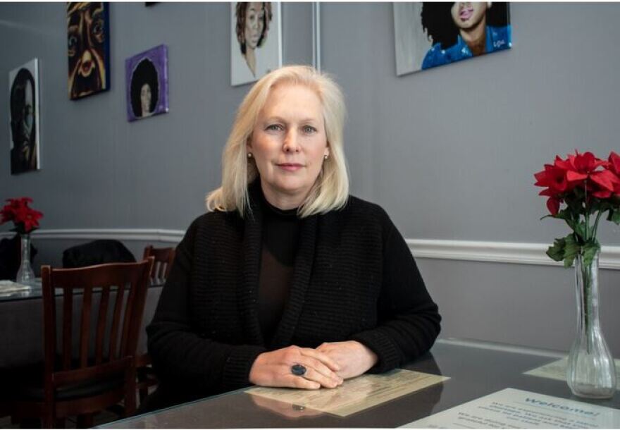 Sen. Kirsten Gillibrand, wearing a black sweater, sits at a desk with a vase of red and green flowers and pictures of women on the wall behind her.