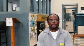 A photo portrait of a Bobby Bostic, who is seated and wearing a gray prison uniform. 