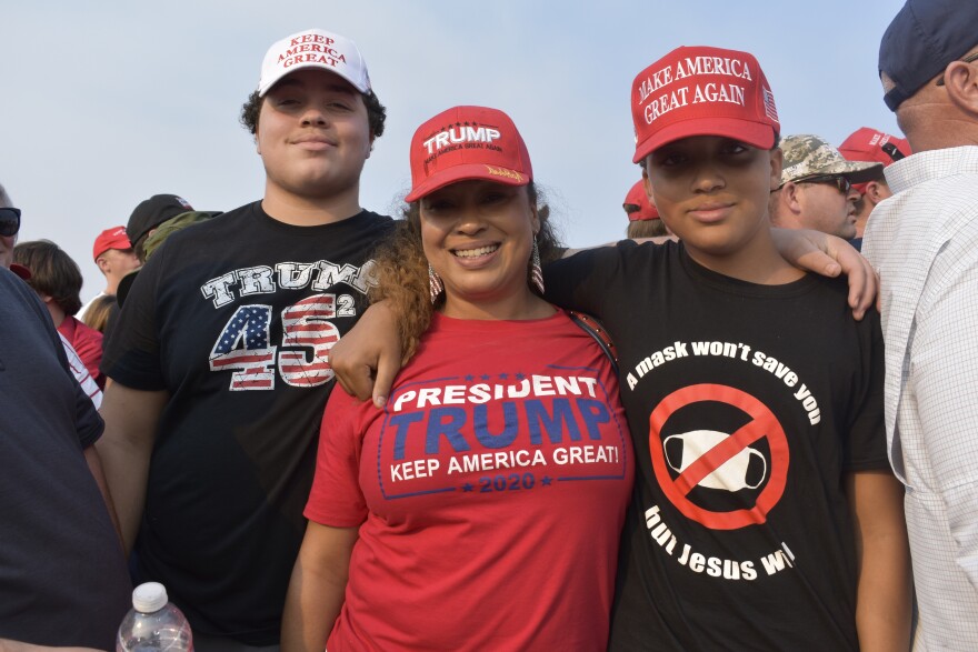 A woman in a Trump shirt with two younger boys on either side of her. One young man has a shirt that says, “A mask won’t save you but Jesus will.”