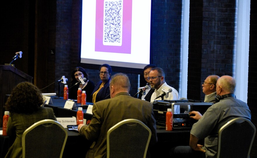  People gathered around a table with a projector screen, engaged in a public forum