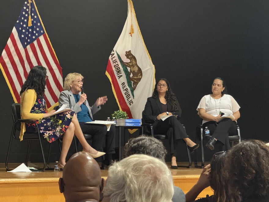 Four people sitting in chairs. The second person from the left is holding a microphone. Behind them are two flags.