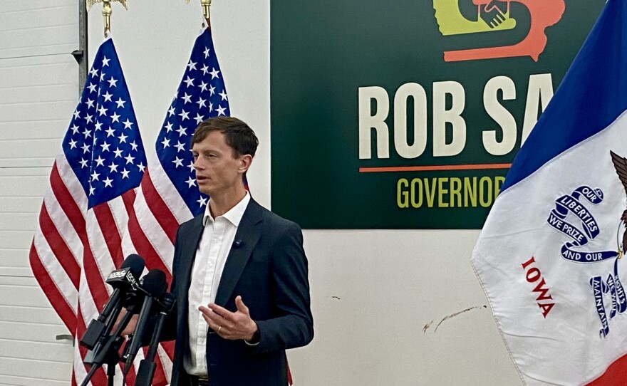 a man speaks into microphones next to American and Iowa flags