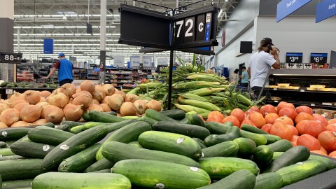 A produce stand at a Walmart store holds cucumbers, tomatoes, onions and corn on the cob. Customers and Walmart workers are visible in the background. 