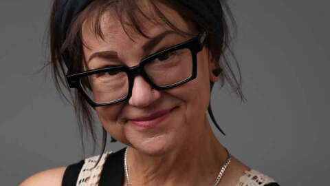 A woman with short brown hair and chunky black glasses smiles at the camera in front of a grey background